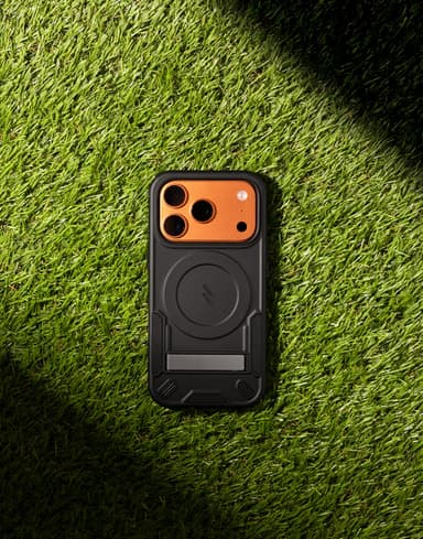 A black ZIZO Transform case with a burnt-orange camera bezel and MagSafe ring, laid flat on green turf under a hard diagonal shadow