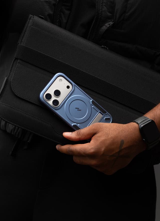 A blue ZIZO Transform MagSafe case cradled in a hand against a black canvas bag, worn with dark streetwear