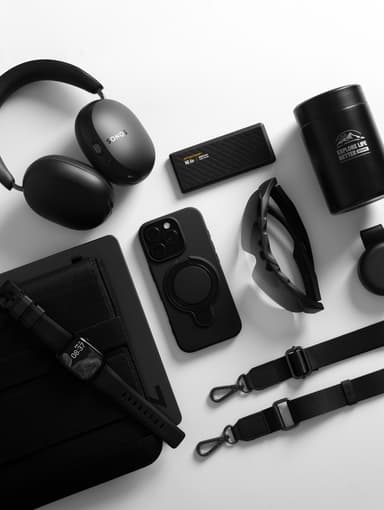 A black ZIZO Revolve case on an iPhone Pro at the center of an all-black everyday-carry flat lay with headphones, a power bank and sunglasses