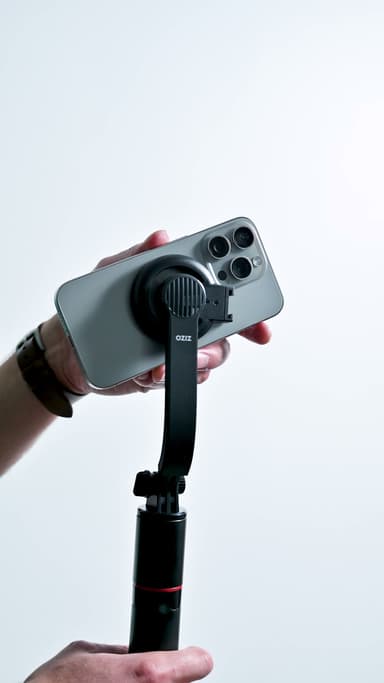 A hand holding an iPhone snapped onto the black MagSafe mount on the ZIZO Gear selfie stick