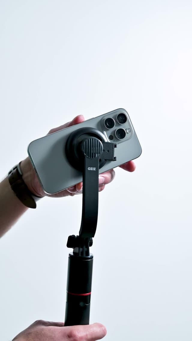 A close-up of an iPhone attached to the black MagSafe mount on the ZIZO Gear selfie stick, on a white studio background