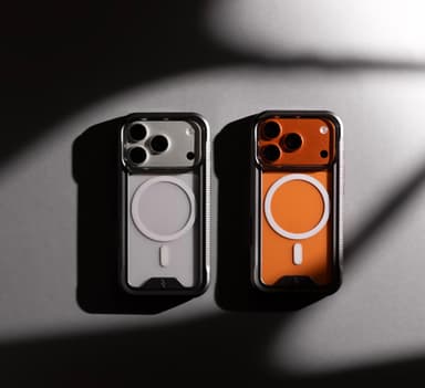 Two ZIZO ION MagSafe cases with black bumpers and clear backs, side by side on a pale surface under hard directional light, one on a silver iPhone 17 Pro and one on a cosmic orange iPhone 17 Pro, both phone finishes reading through the clear back