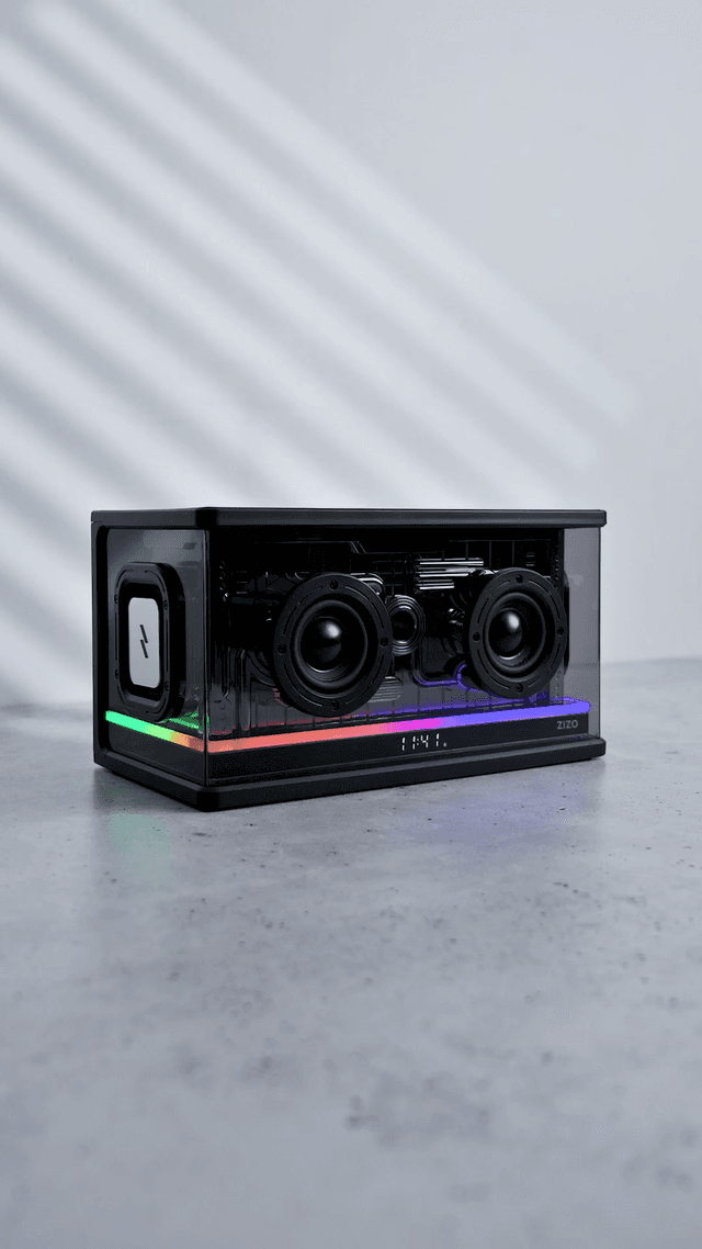 The ZIZO Retro Crystal speaker on a gray studio floor, twin drivers behind its transparent shell and the RGB bar lit across the front