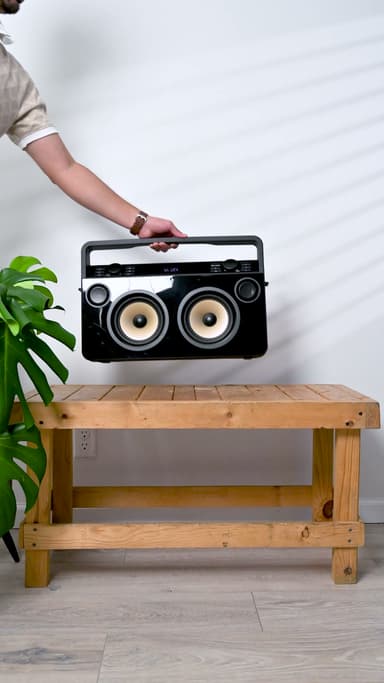 The ZIZO Retro boombox in black with twin cream woofers, held by its handle above a light wood bench beside a monstera plant