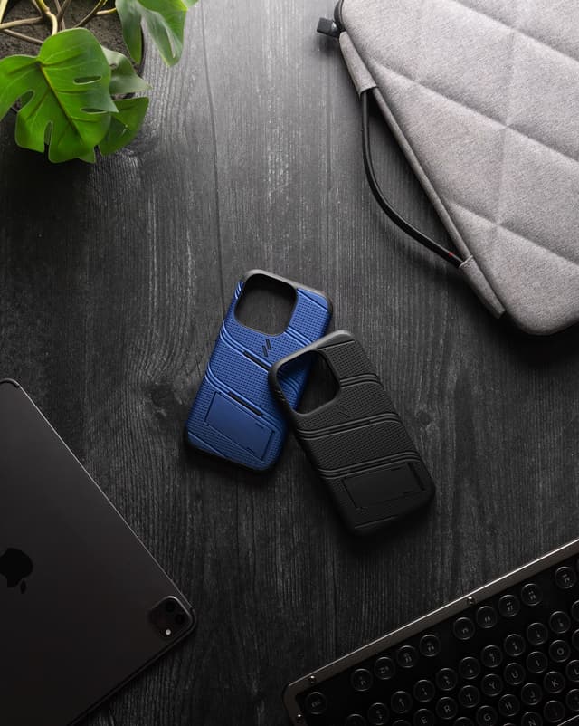 ZIZO Bolt cases in navy blue and black leaning on a dark wood desk with a plant, keyboard, and laptop sleeve