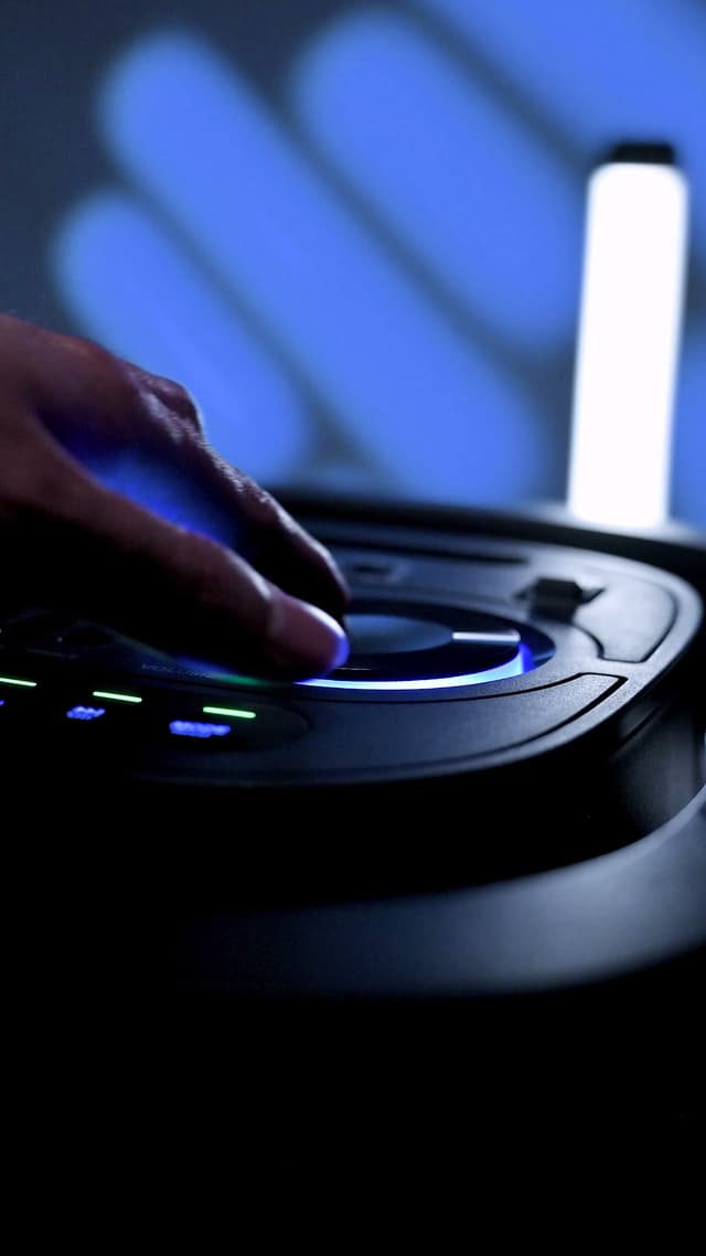 A finger pressing a ring-lit control on the ZIZO Boom speaker's panel in a dark blue-lit room