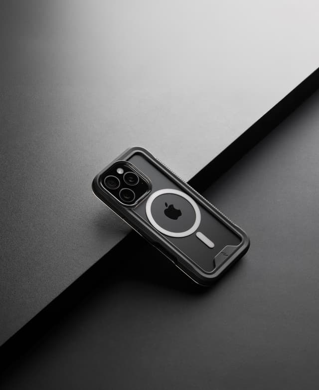 A ZIZO ION MagSafe case with a smoke bumper and clear back on a dark phone, resting on a dark two-tone surface under hard directional light, the magnet ring and textured side rail catching the light