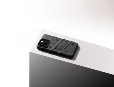 A black ZIZO BOLT rugged case on an iPhone, resting diagonally on the edge of a white pedestal, its dot-grid textured back and built-in kickstand raked by studio light