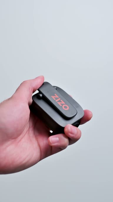 The black ZIZO Sonic Clip speaker with its orange ZIZO mark, its clip held open in a hand against a light gray ground