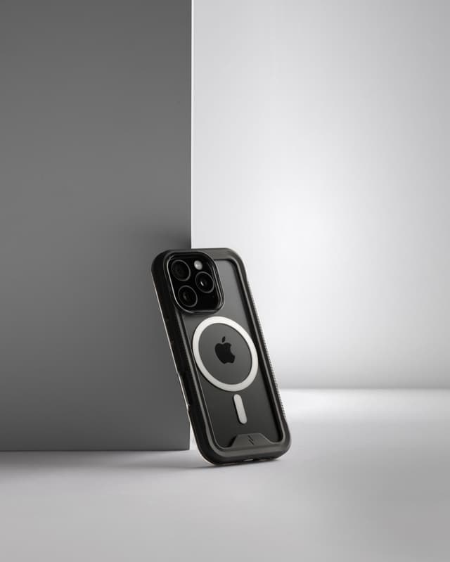 A ZIZO ION MagSafe case with a smoke bumper and clear back, standing upright against a two-tone gray and white studio backdrop, its raised bezel edge and textured side rail in clean silhouette