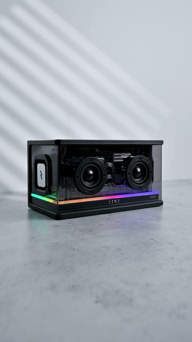 The ZIZO Retro Crystal speaker turning on a gray studio floor, its RGB light bar cycling color and the drivers visible through the transparent shell