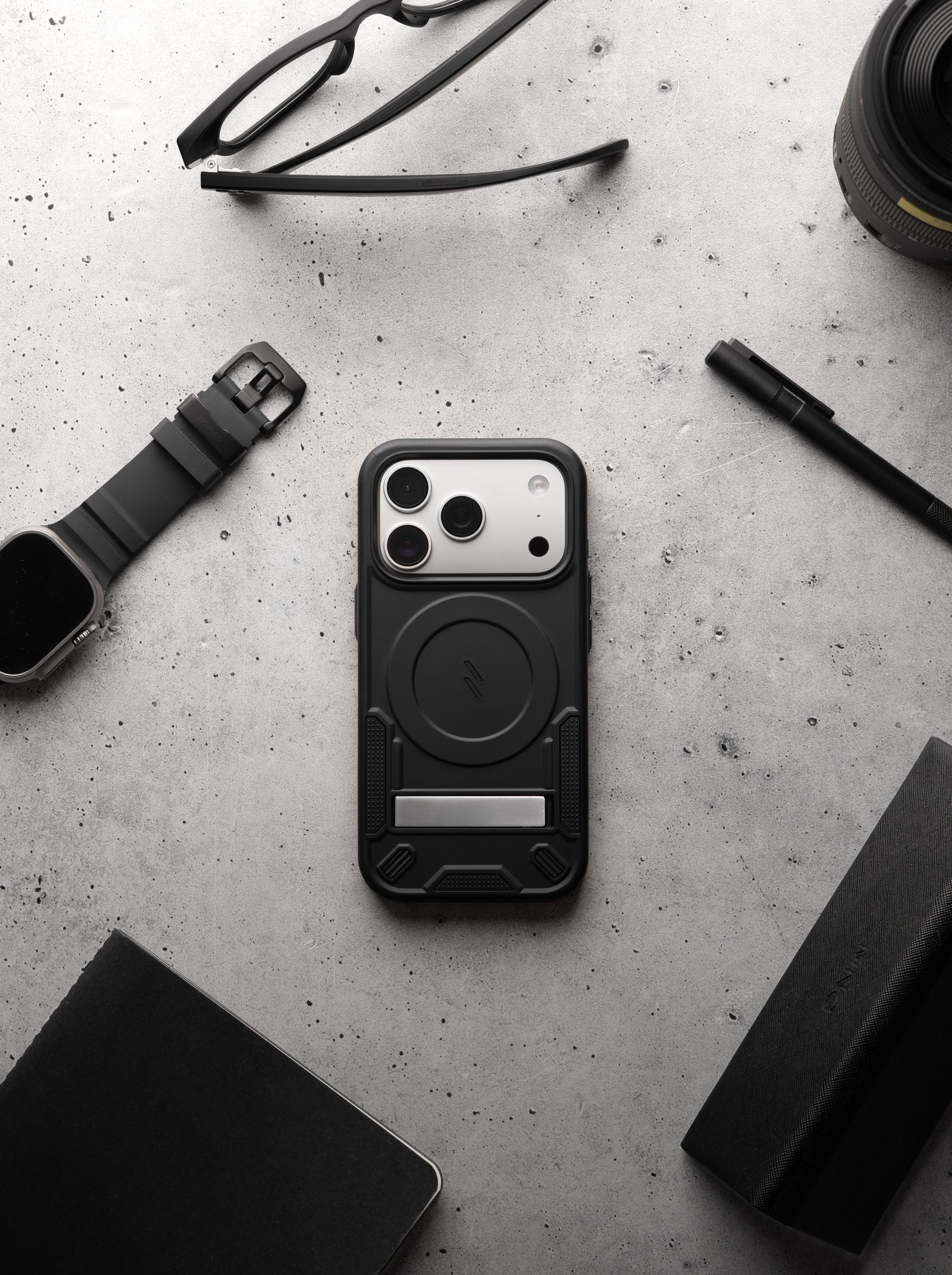 A black ZIZO Transform case on an iPhone Pro staged among glasses, a smartwatch, a pen, a notebook and a camera lens on concrete