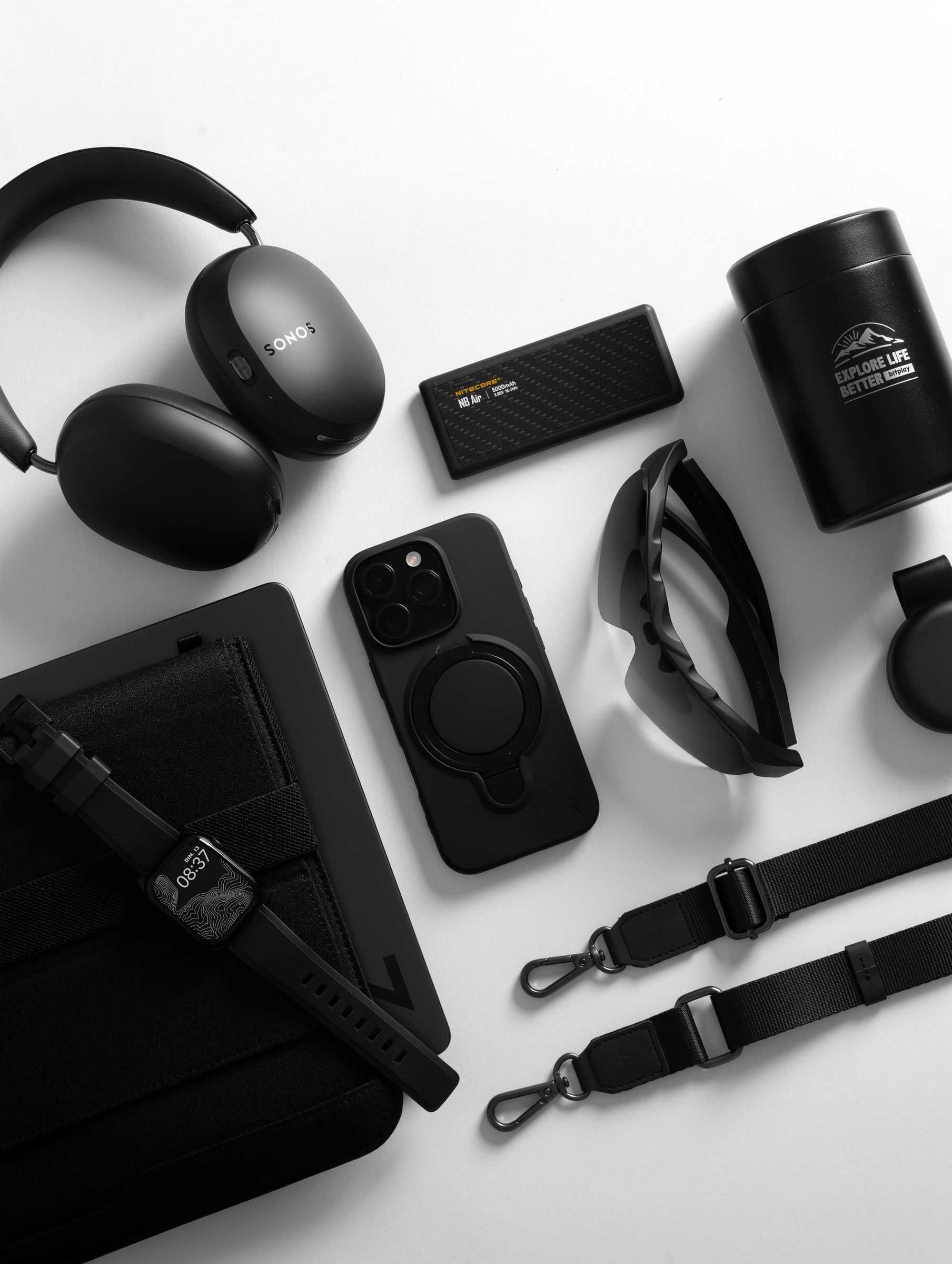 A black ZIZO Revolve case on an iPhone Pro at the center of an all-black everyday-carry flat lay with headphones, a power bank and sunglasses
