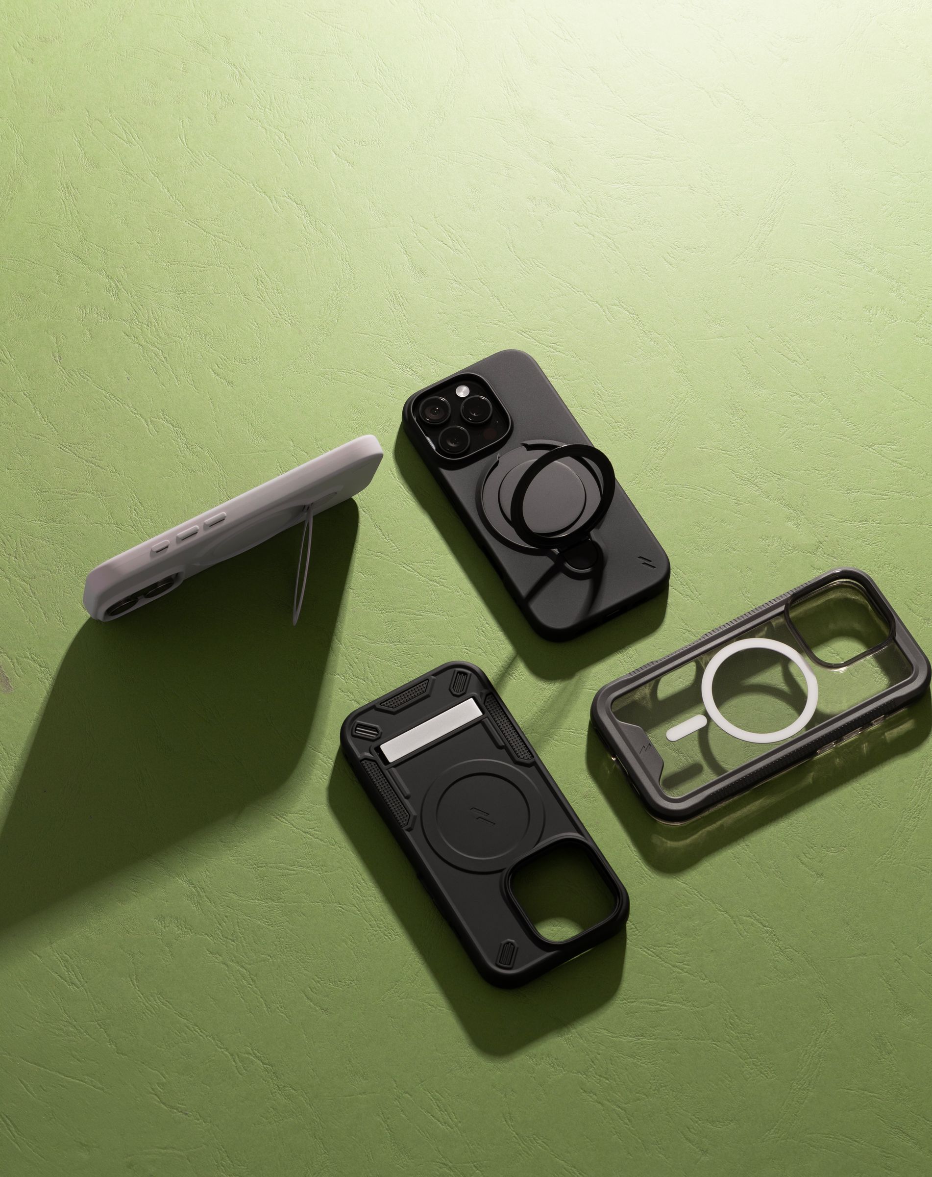 Four ZIZO cases arranged on a textured green surface, including a ring-stand case, a card-holder case with a kickstand, and a clear ION MagSafe case at the right