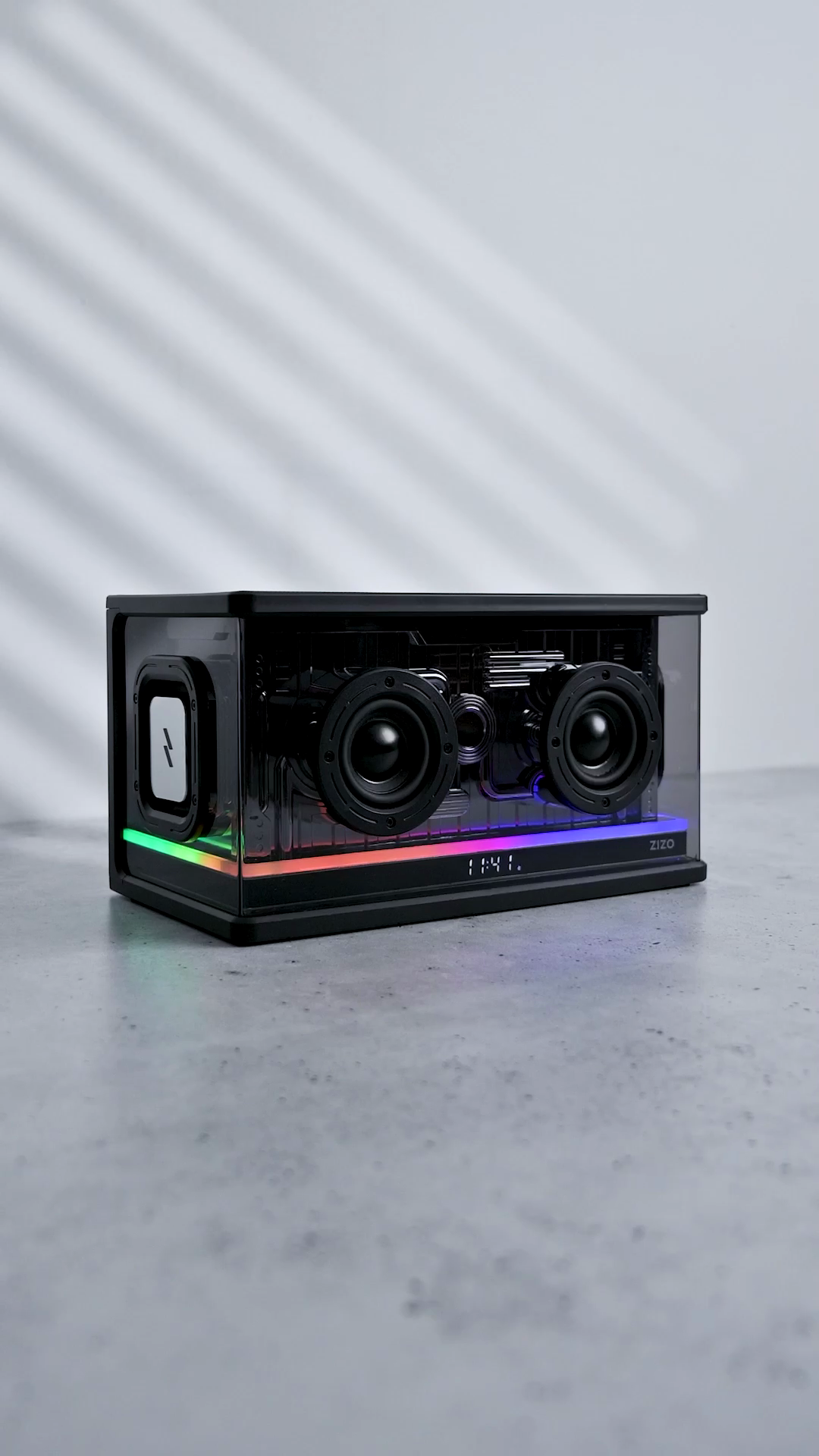 The ZIZO Retro Crystal speaker on a gray studio floor, twin drivers behind its transparent shell and the RGB bar lit across the front