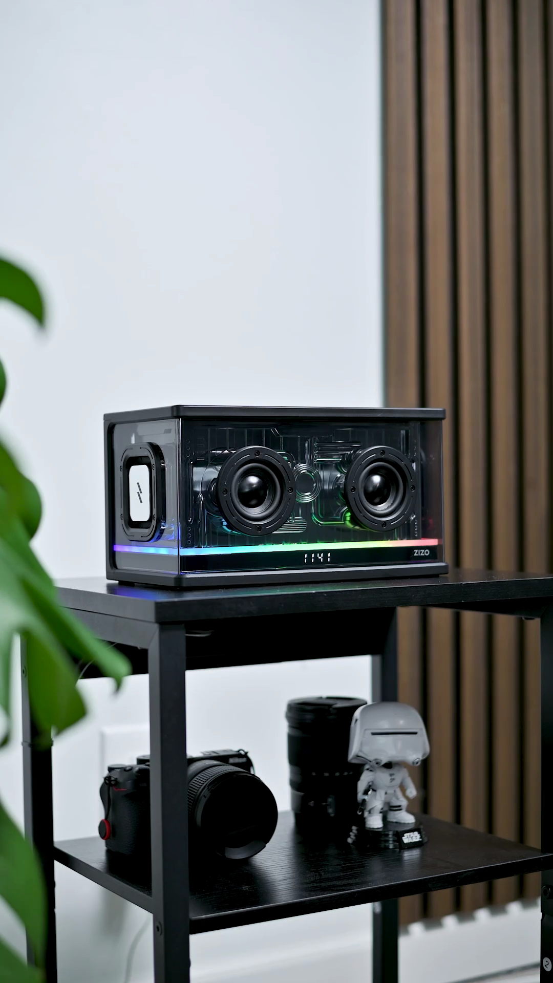 The ZIZO Retro Crystal speaker on a black shelf beside cameras and a plant, its RGB bar lit in rainbow