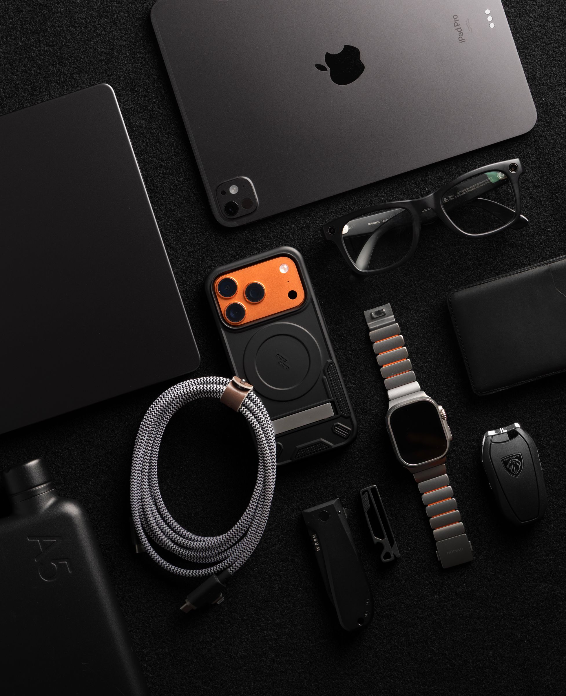 A black ZIZO Transform case with an orange camera plate laid into an all-black everyday-carry flat lay with an iPad Pro, watch and wallet