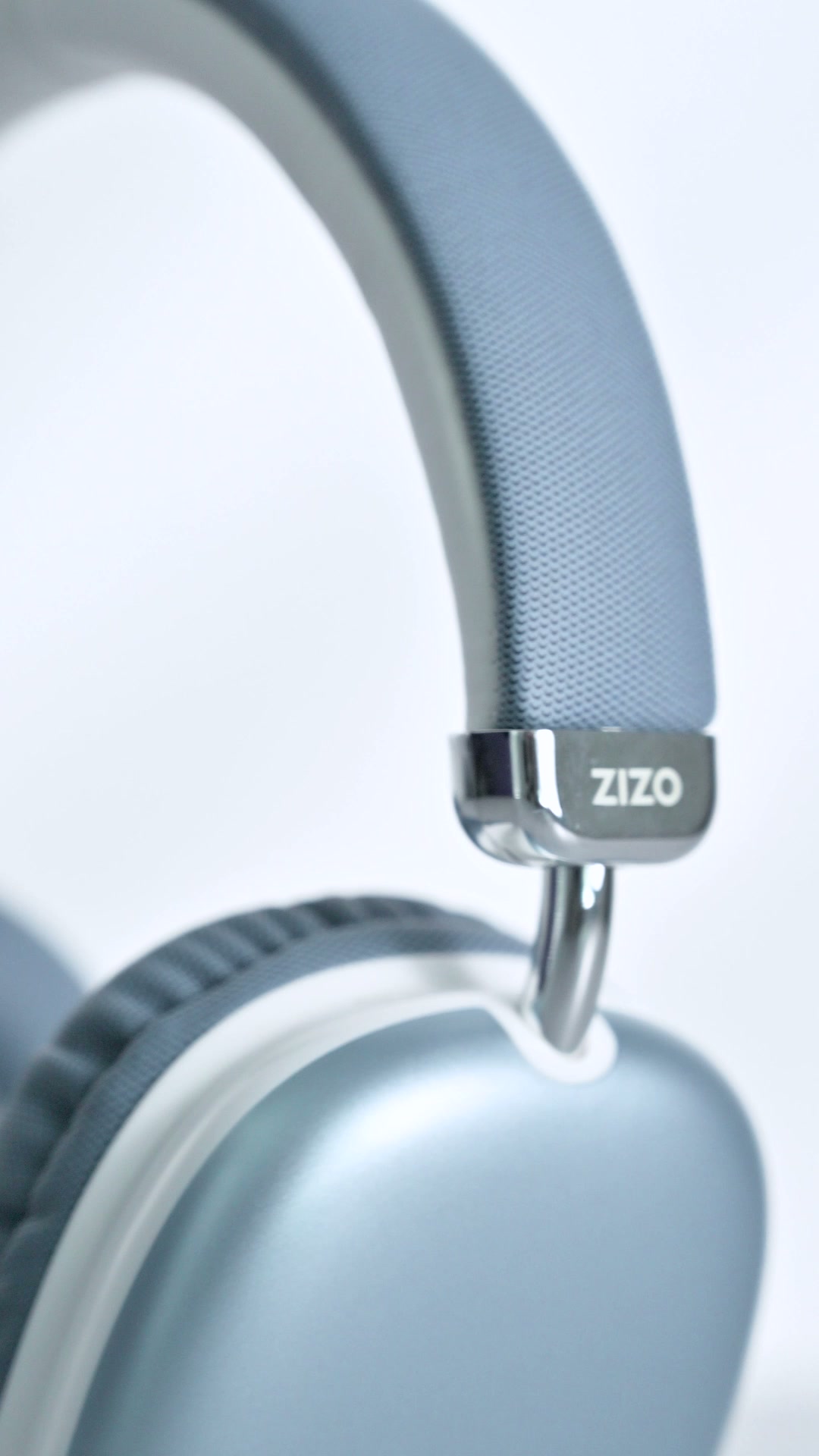 A close view of the polished metal hinge on a pale ZIZO Tempo Pro headphone, the ZIZO mark on the plate and the woven fabric band above it