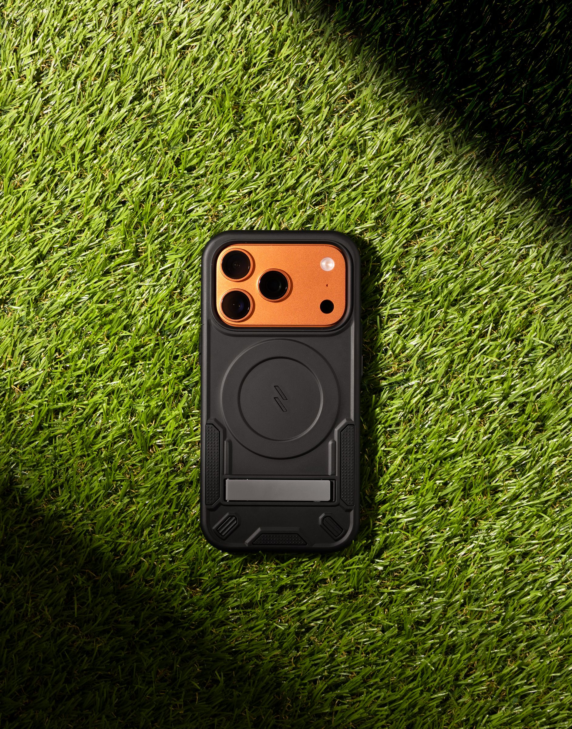 A black ZIZO Transform case with a burnt-orange camera bezel and MagSafe ring, laid flat on green turf under a hard diagonal shadow